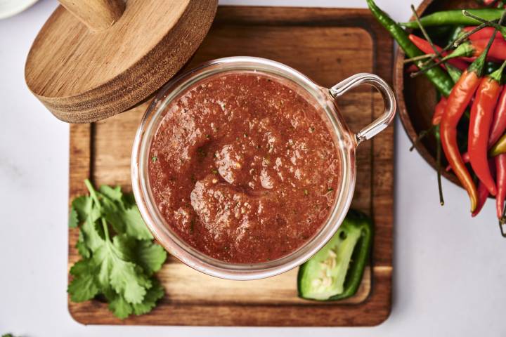 Homemade Salsa Roja A glass jar filled with chunky salsa rests on a wooden board, surrounded by fresh cilantro and jalapeños, with vibrant chili peppers nearby.