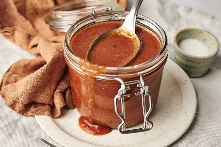 Shortcut Enchilada Sauce A glass jar filled with homemade enchilada sauce, with a spoon resting inside, placed on a ceramic plate with a rustic cloth and a small bowl of salt in the background.