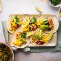 Salmon Tacos with Corn Salsa Salmon tacos with corn salsa, mango, fresh avocado slices, and lime wedges on a tray.