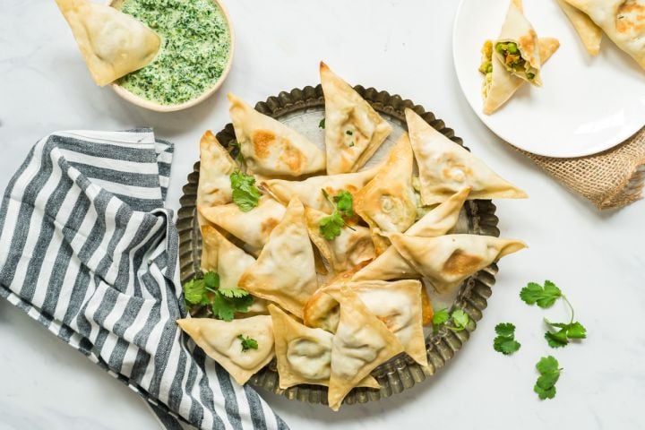 Shortcut Baked Samosa Baked samosa filled with potato, cauliflower, and peas on a plate with cilantro and mint dipping sauce.