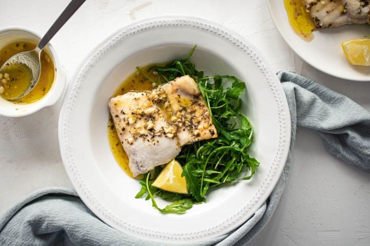 Easy Baked Cod Easy baked cod with a lemon, garlic, and butter sauce on a white plate with mixed greens.