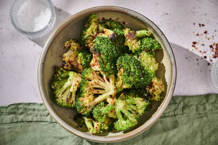 Garlic Grilled Broccoli | How to Grill Broccoli A bowl of roasted broccoli with crispy edges, sprinkled with red pepper flakes, sits on a light surface beside a salt dish.