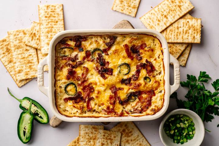 High Protein Jalapeño Popper Chicken Dip A creamy baked dish topped with bacon and jalapeños is centered in a rectangular dish. Surrounding the dish are several crackers, sliced jalapeño and fresh parsley, with a small bowl of chopped green onions.