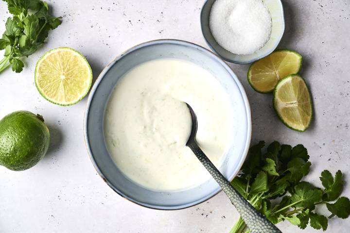 Lime Crema Recipe A bowl of creamy lime crema with a spoon, surrounded by fresh cilantro, lime halves, and a dish of sugar.