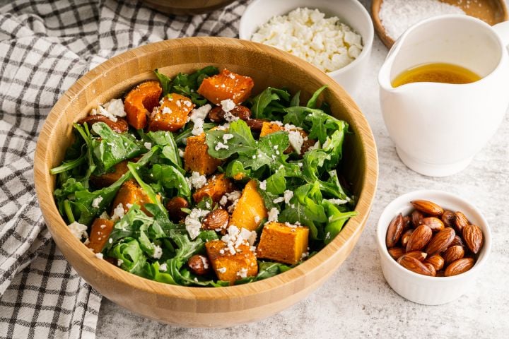 Roasted Butternut Squash Salad Roasted butternut squash salad with arugula, roasted squash, almonds, feta cheese, and orange vinaigrette in a wooden bowl.