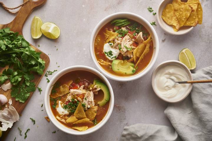 Slow Cooker Soup Recipes for Busy Weeknights Two bowls of slow cooker chicken fajita soup topped with crispy tortilla chips, avocado slices, sour cream, and fresh cilantro, served with lime wedges and garnishes on a neutral background.