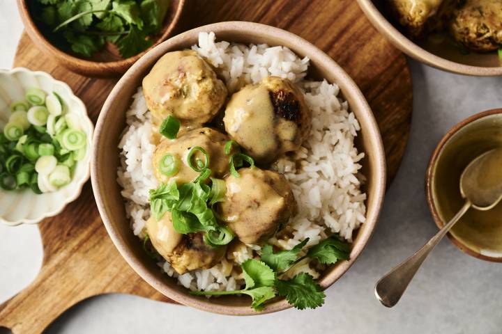 Slow Cooker Thai Curry Chicken Meatballs A bowl of white rice topped with Thai curry chicken meatballs, coated in a creamy sauce and garnished with fresh cilantro and sliced green onions.