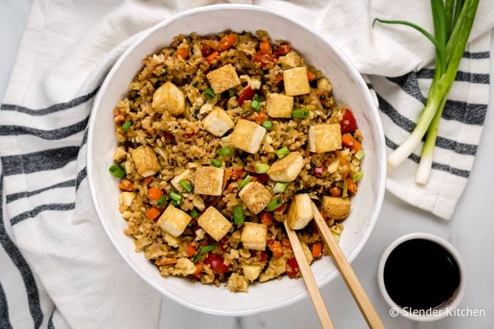 Tofu Cauliflower Fried Rice Tofu cauliflower fried rice with assorted vegetables, green onions, and soy sauce in a bowl with chopsticks.