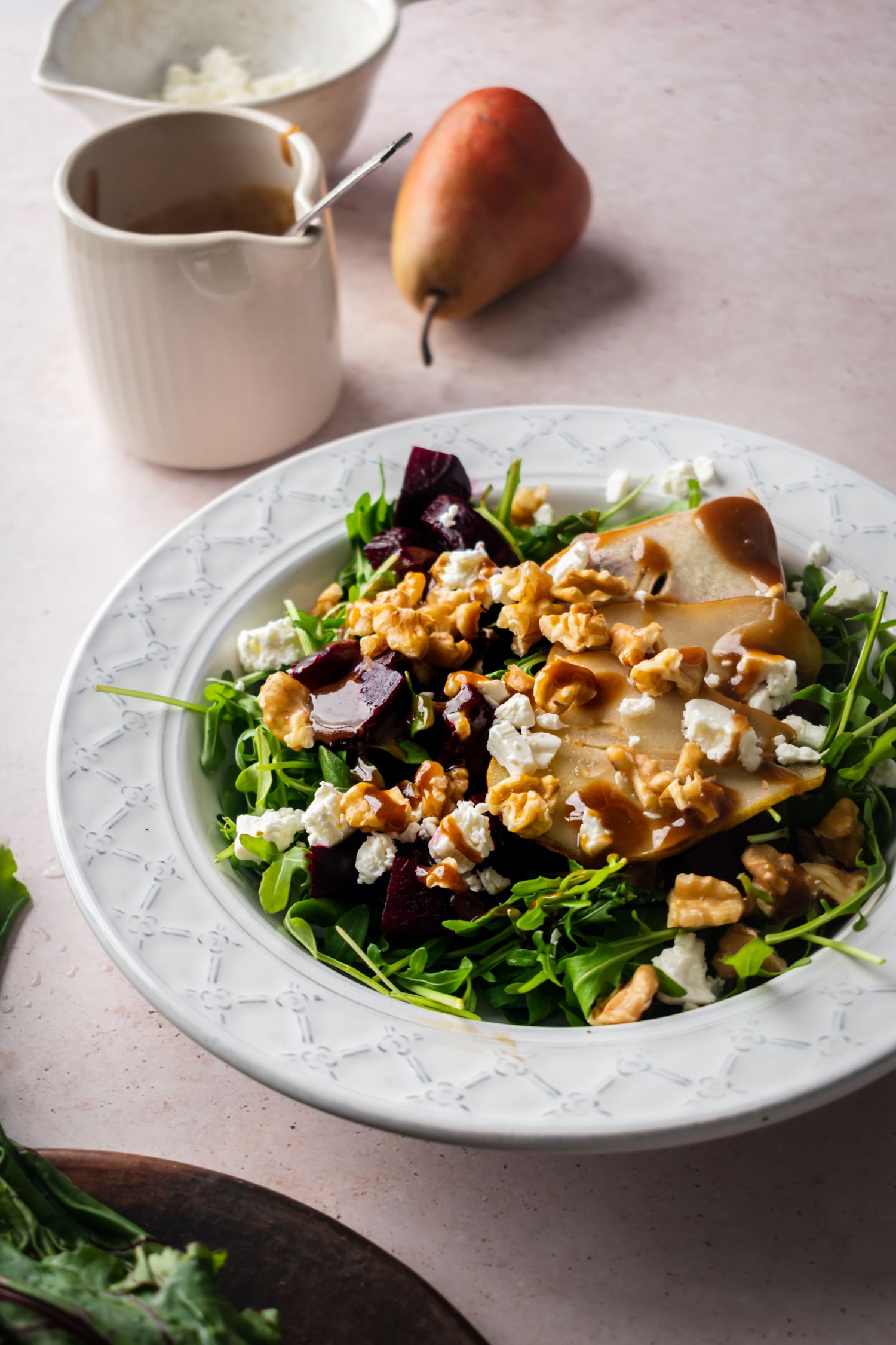 Baked beet salad with walnuts, goat cheese, sliced pears, and arugula in a bowl with balsamic dressing on the side.