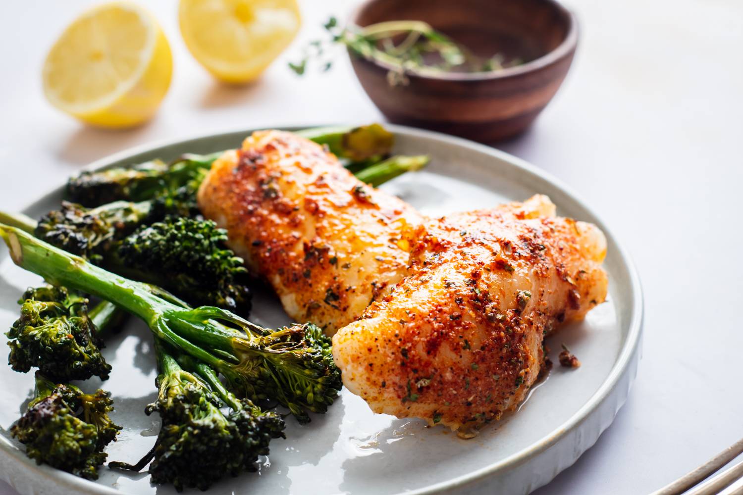 Two seasoned fish fillets on a plate, accompanied by roasted broccolini and sliced lemons. A small bowl of herbs is nearby.
