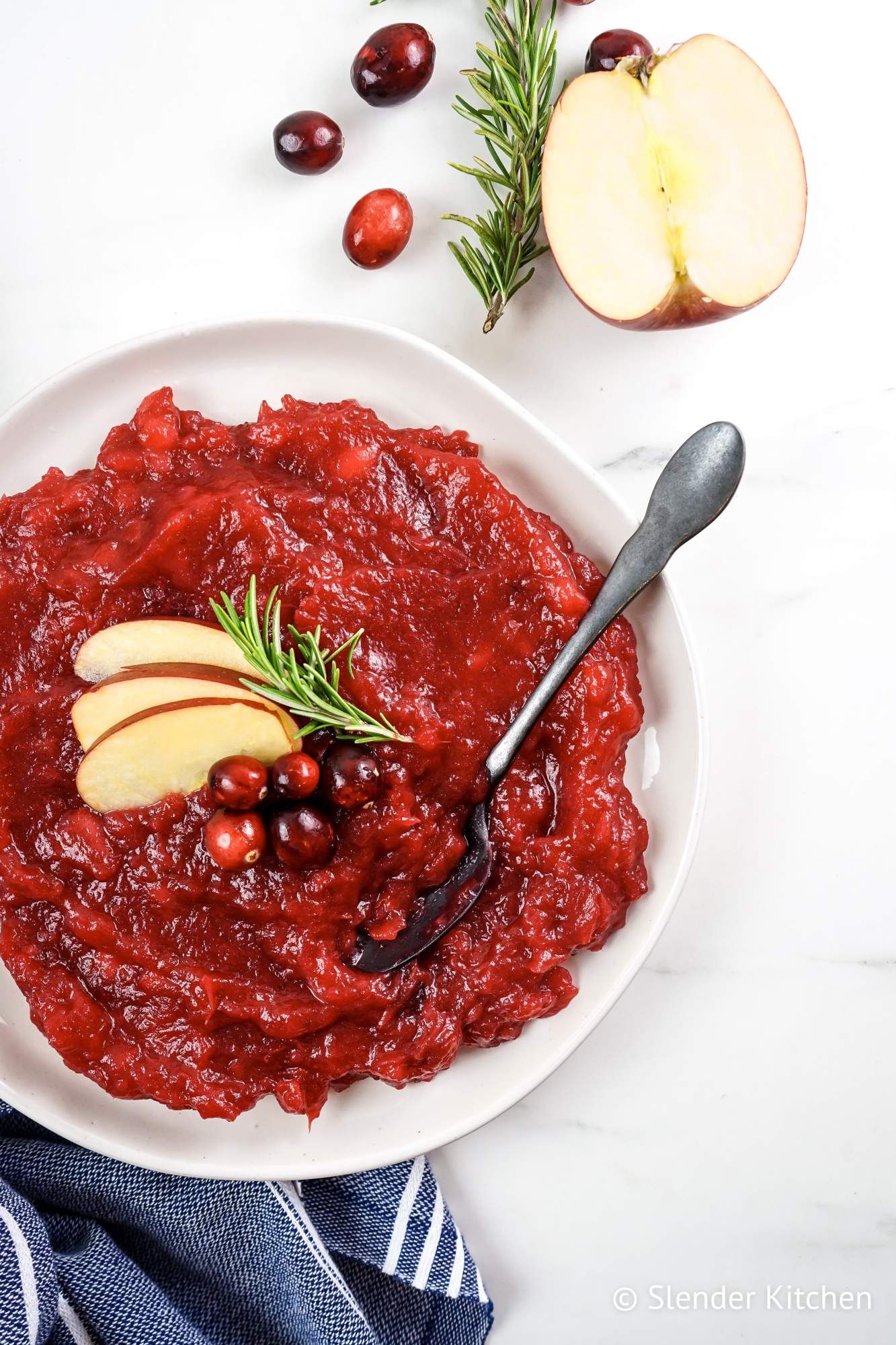 Apple and cranberry sauce served in a bowl with a spoon, fresh cranberries, apple slices, and rosemary,