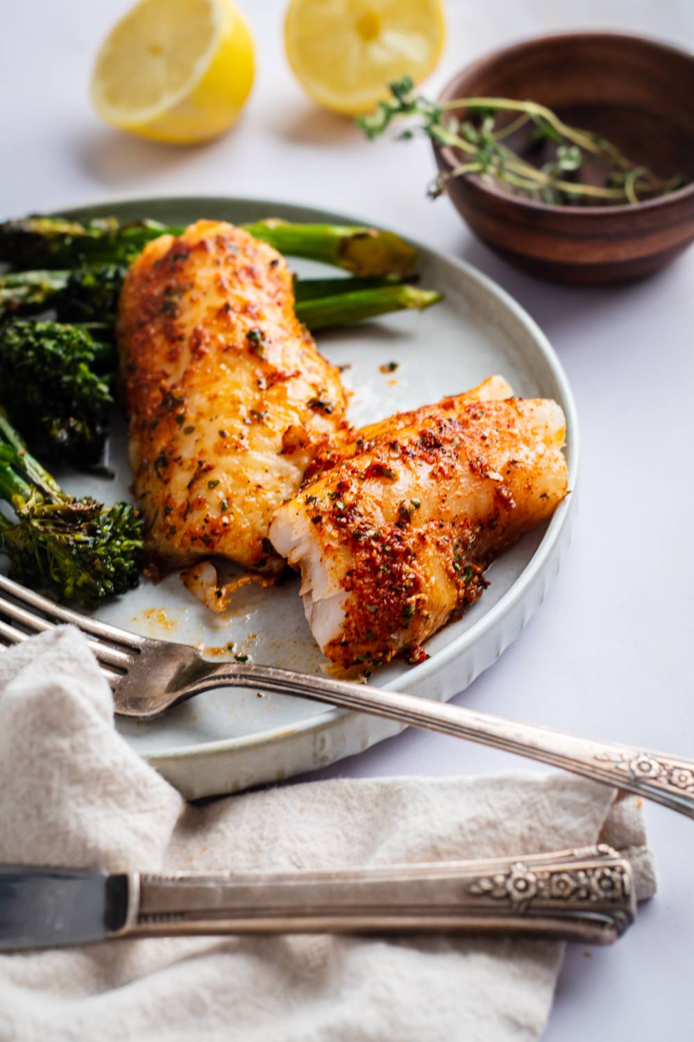 A plate of seasoned fish fillets with roasted broccoli, garnished with thyme, accompanied by lemon halves and vintage silverware.