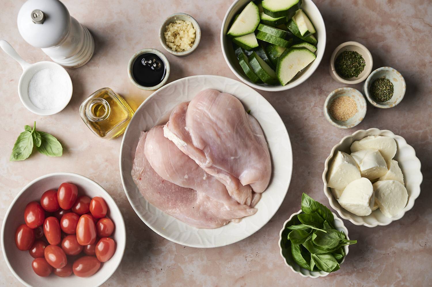 A plate of raw chicken breasts, grape tomatoes, zucchini slices, mozzarella, and fresh basil with seasonings ready for cooking.