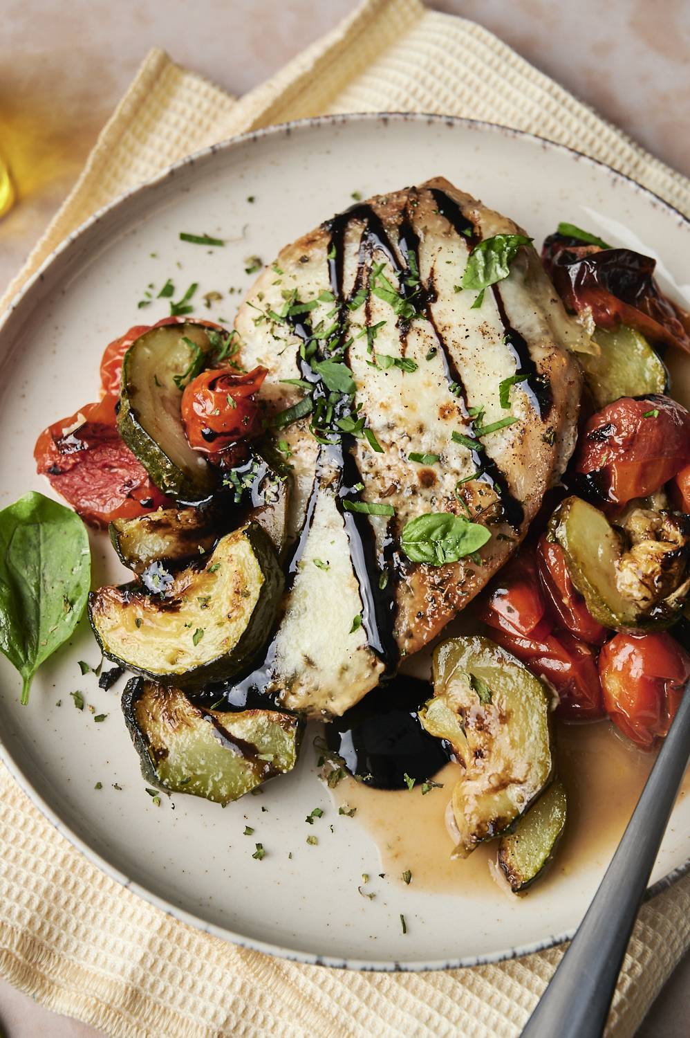 A plate with a serving of caprese chicken, topped with balsamic glaze and fresh basil, served with roasted zucchini and tomatoes.