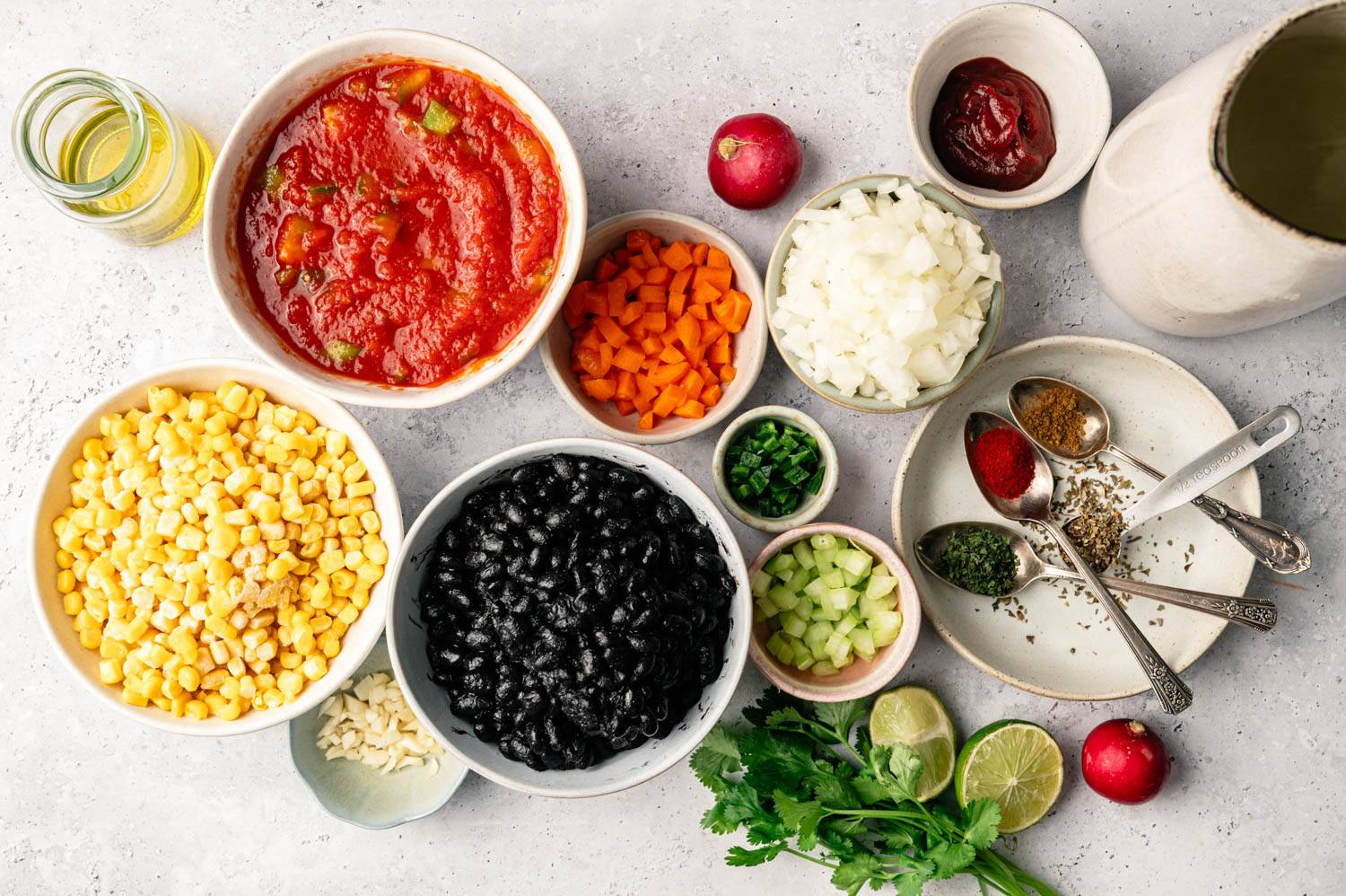 Colorful ingredients for a vibrant recipe are neatly arranged on a light surface. Includes bowls of tomatoes, corn, black beans, spices, and fresh herbs.