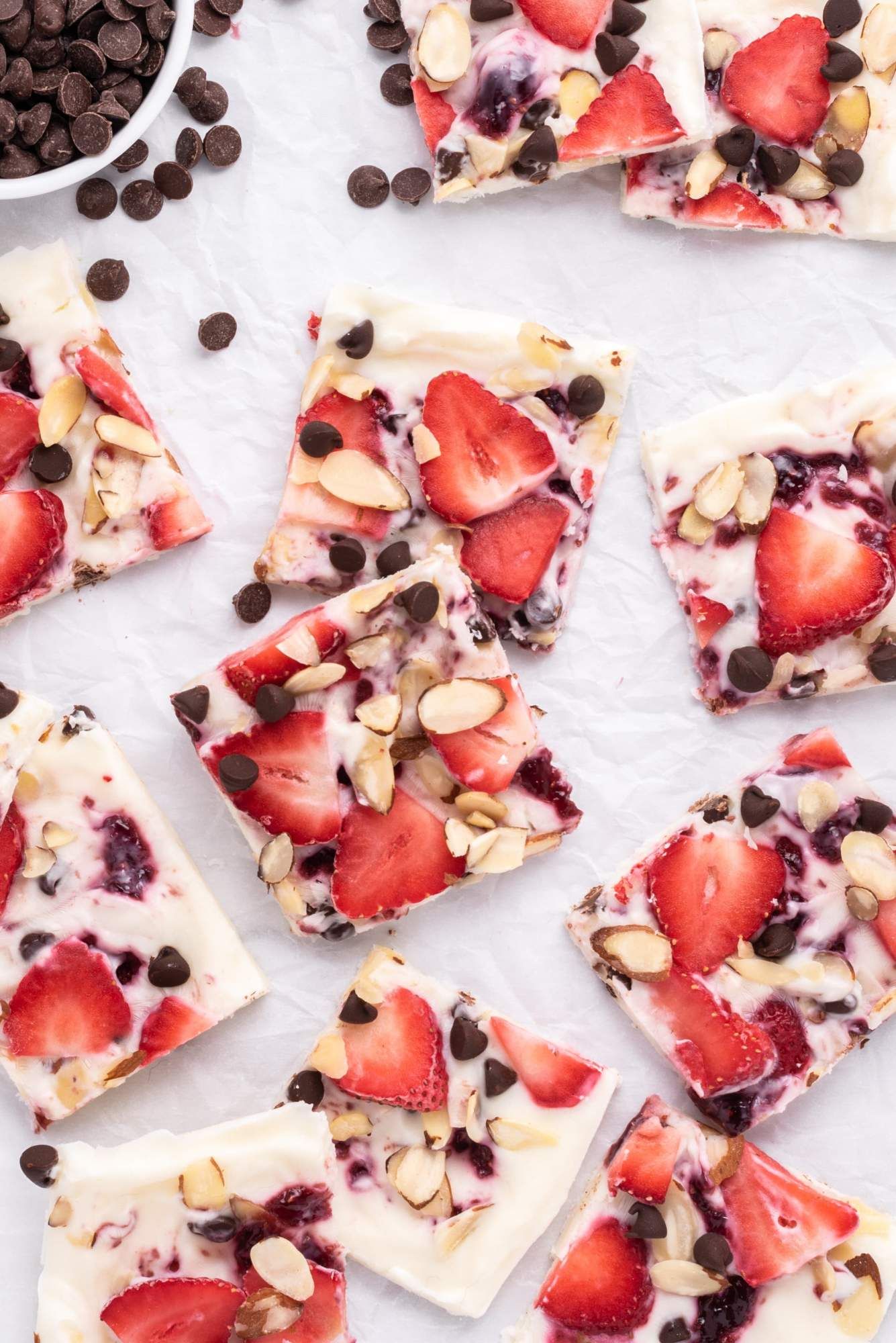 Frozen yogurt bark with sliced strawberries, chocolate chips, and almonds broken into pieces on parchment paper.