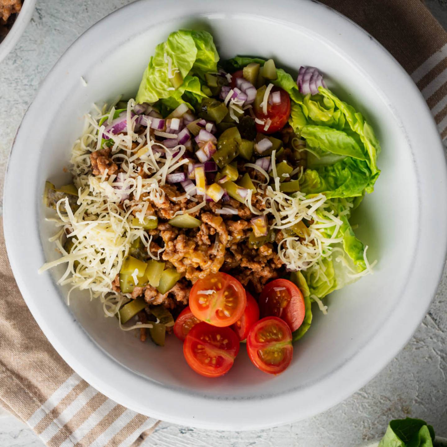 Cheeseburger salad with ground beef, pickles, tomatoes, lettuce, and shredded cheddar cheese in a bowl.