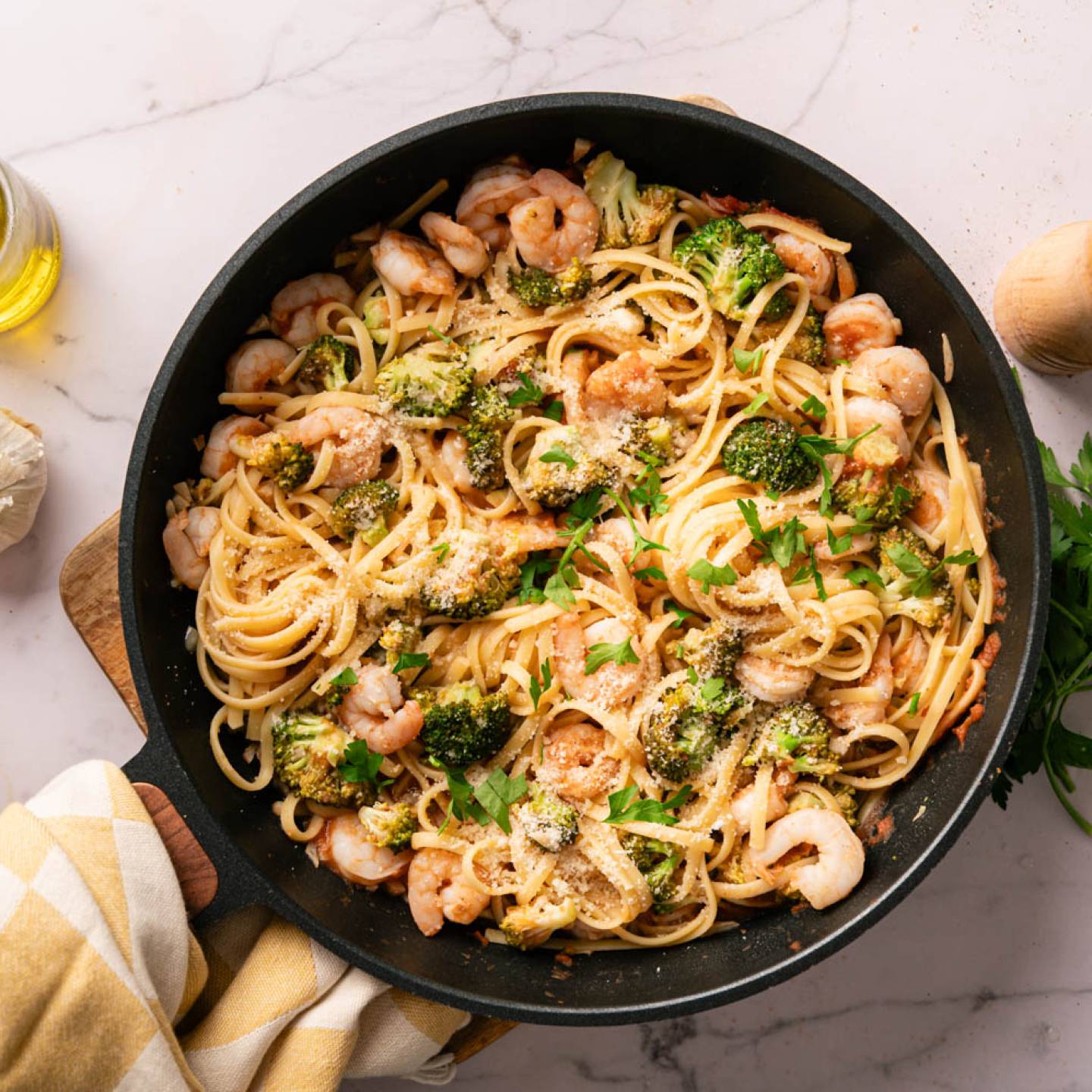 A skillet of shrimp and broccoli linguine, garnished with parsley and Parmesan. Surrounding are garlic cloves, olive oil, and a pepper mill on a marble surface.