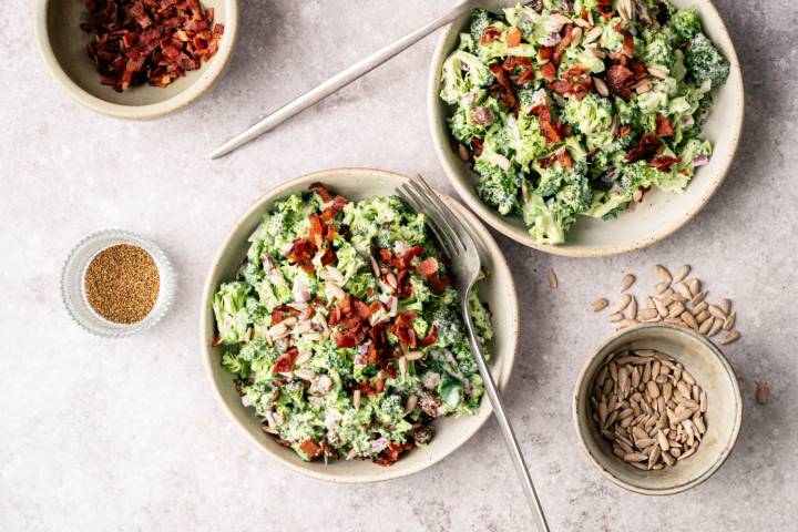 Two bowls of creamy broccoli salad topped with bacon and seeds are on a marble surface. Nearby are small bowls of bacon bits, sunflower seeds, and a fork.