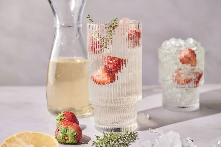 A tall glass of white wine spritzer garnished with strawberries and thyme sits next to a carafe of wine.