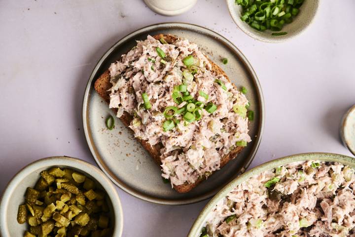 A top-down shot of an open-faced dill pickle tuna salad sandwich, with bowls of the salad, pickles, and green onions on the side.