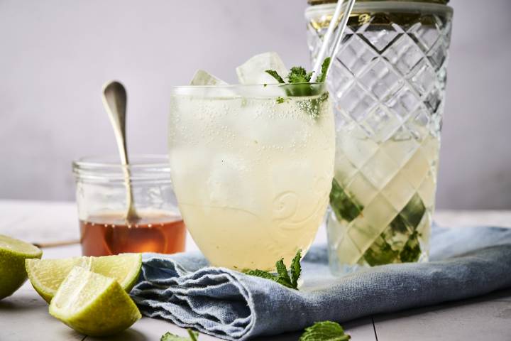 A refreshing drink with mint and ice, next to a jar of honey and lime wedges on a blue napkin.