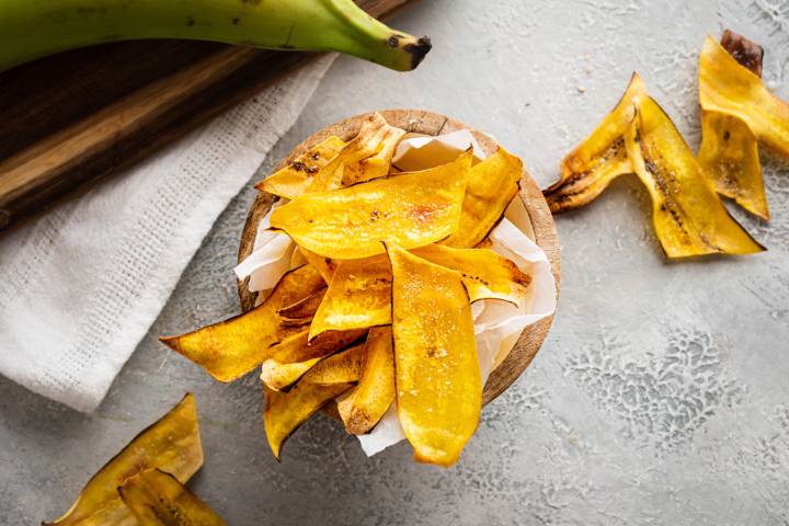Wooden bowl filled with golden homemade plantain chips, sprinkled with salt, surrounded by extra chips and a whole green plantain on a textured gray surface.