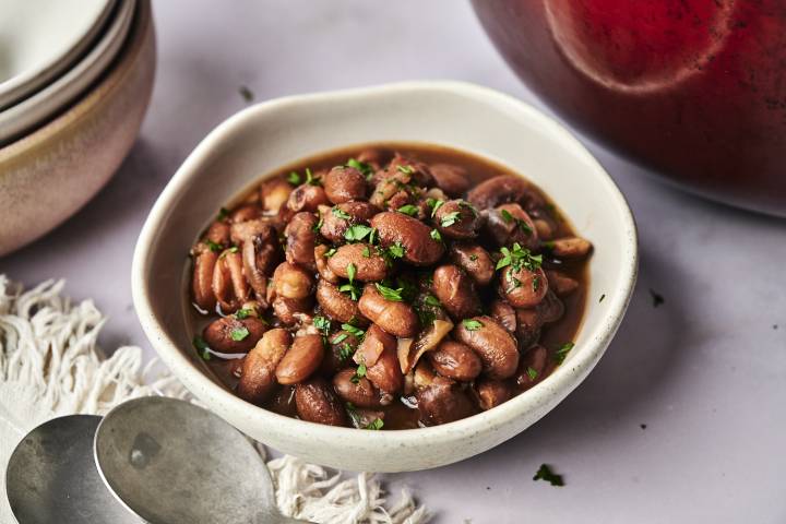 A bowl of savory, cooked beans garnished with fresh parsley, accompanied by shiny metal spoons and delicate dishware, set on a table.