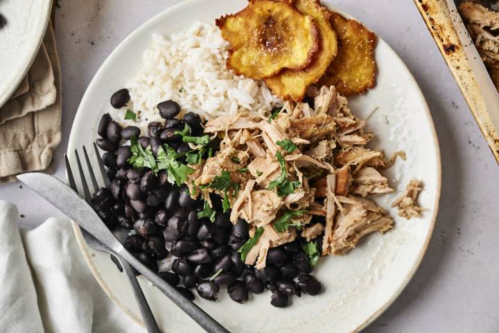 Shredded garlic Cuban pork served with black beans, rice, and crispy plantains on a white plate. The pork is topped with fresh cilantro, and there is a fork and knife on the side. 