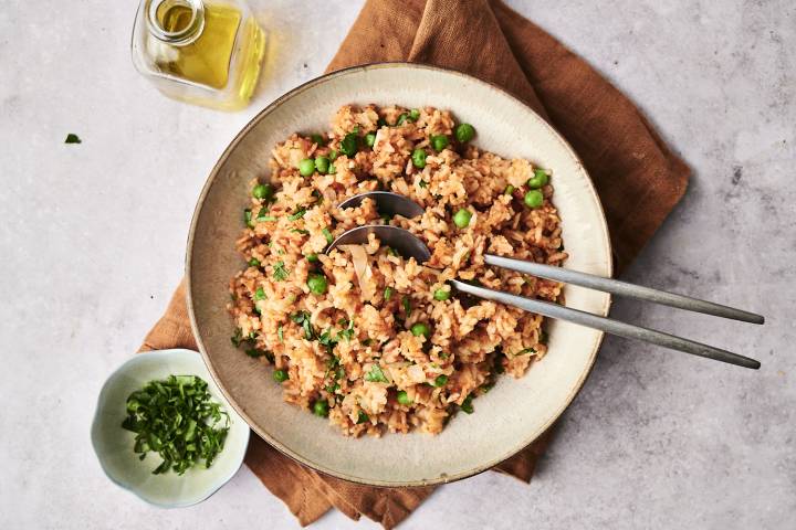 Overhead view of a bowl of slow cooker Mexican rice with green peas and chopped cilantro, served with two metal spoons on a brown napkin, next to a small bowl of herbs and a glass bottle of olive oil.  