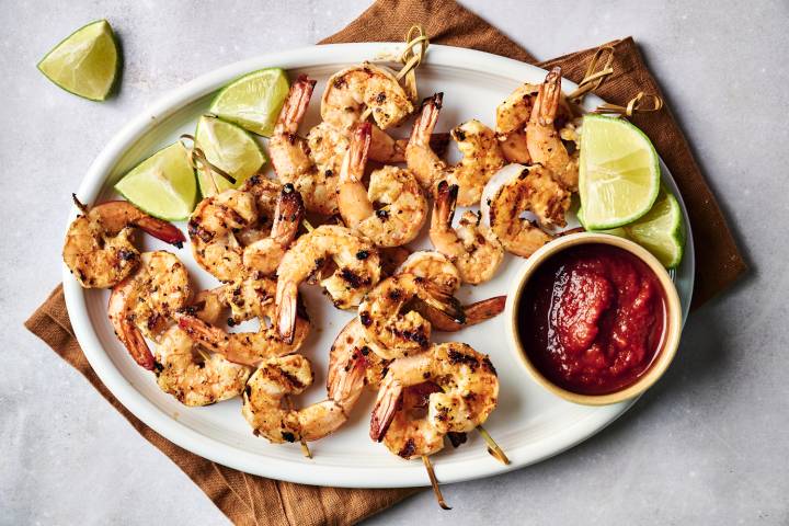 Grilled shrimp skewers served with lime wedges and a bowl of dipping sauce on a white platter over a brown cloth.
