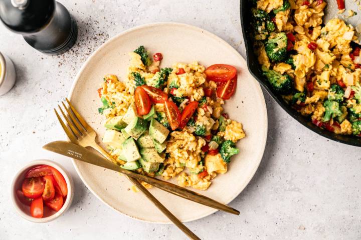 A plate of scrambled eggs mixed with broccoli and red peppers, topped with avocado and cherry tomatoes. A fork and knife rest beside it. A bowl of tomatoes and a skillet are nearby. The setting is bright and appetizing.