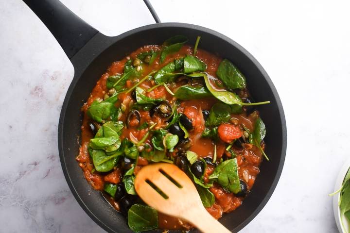 Spinach, black olives, and capers being stirred into tomato sauce.