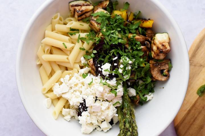 Cooked pasta, feta cheese, fresh herbs, and grilled vegetables in a bowl for pasta salad.