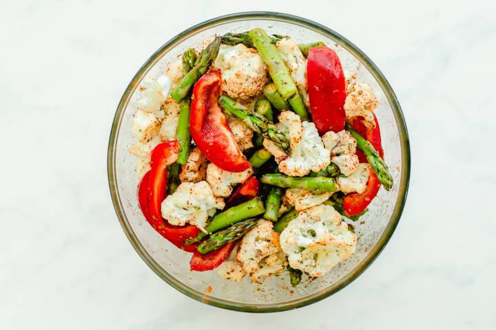 Cauliflower, asparagus, and red pepper slices in a bowl with olive oil and spices.