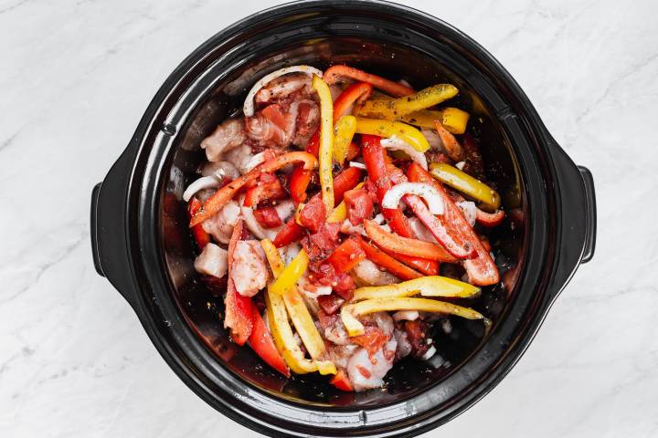 Slow cooker filled with chicken breast, red peppers, onions, tomatoes, and herbs.