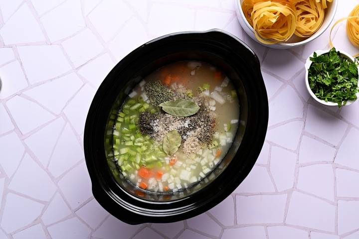 Celery, carrots, onion, garlic, broth, and spices being added to a slow cooker with turkey.