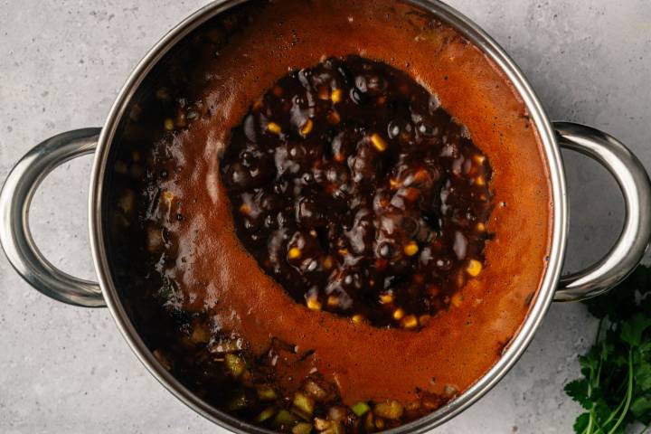 A stainless steel pot filled with rich, bubbling black bean mixture, corn kernels, and visible diced vegetables on a light gray countertop.