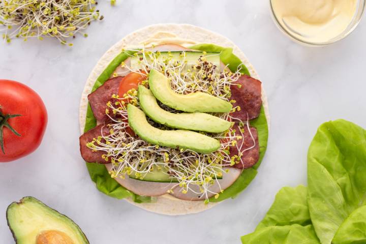 Turkey wrap being assembled with deli turkey, bacon, lettuce, tomato, avocado, and sprouts.