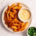 Plate of crispy golden fries with a creamy dipping sauce on the side. A small bowl of fresh green parsley is nearby on a light, textured surface.