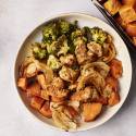A serving of sheet pan herb chicken and sweet potatoes plated with roasted broccoli, caramelized onions, and seasoned sweet potato chunks. The plate is set on a light background with a portion of the sheet pan visible beside it.