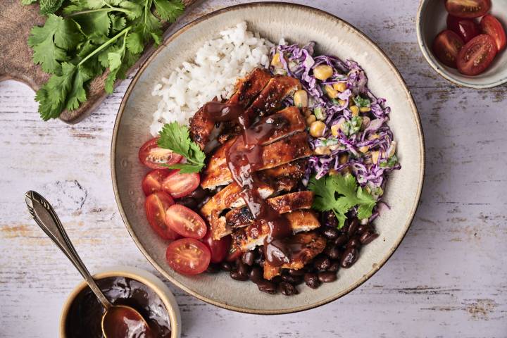 A colorful bowl featuring grilled chicken, rice, black beans, cherry tomatoes, slaw, and herbs, drizzled with barbecue sauce.