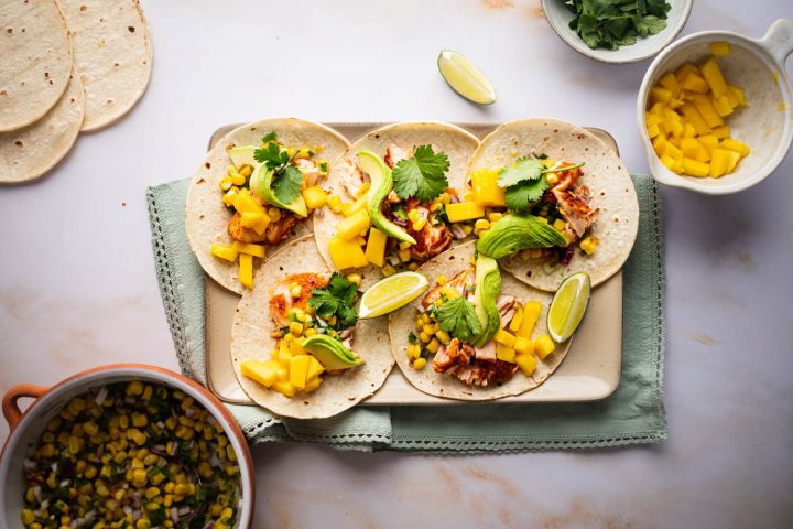 Salmon tacos with corn salsa, mango, fresh avocado slices, and lime wedges on a tray. 