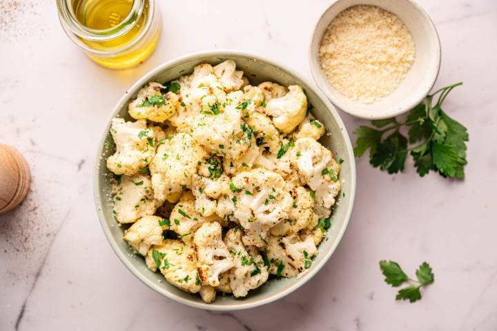 A bowl of roasted cauliflower florets garnished with chopped parsley on a marble surface. Nearby are a bowl of grated cheese and a jar of olive oil.