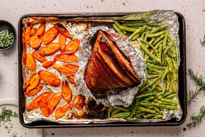 A roasted ham surrounded by glazed carrots and green beans on a foil-lined baking sheet.