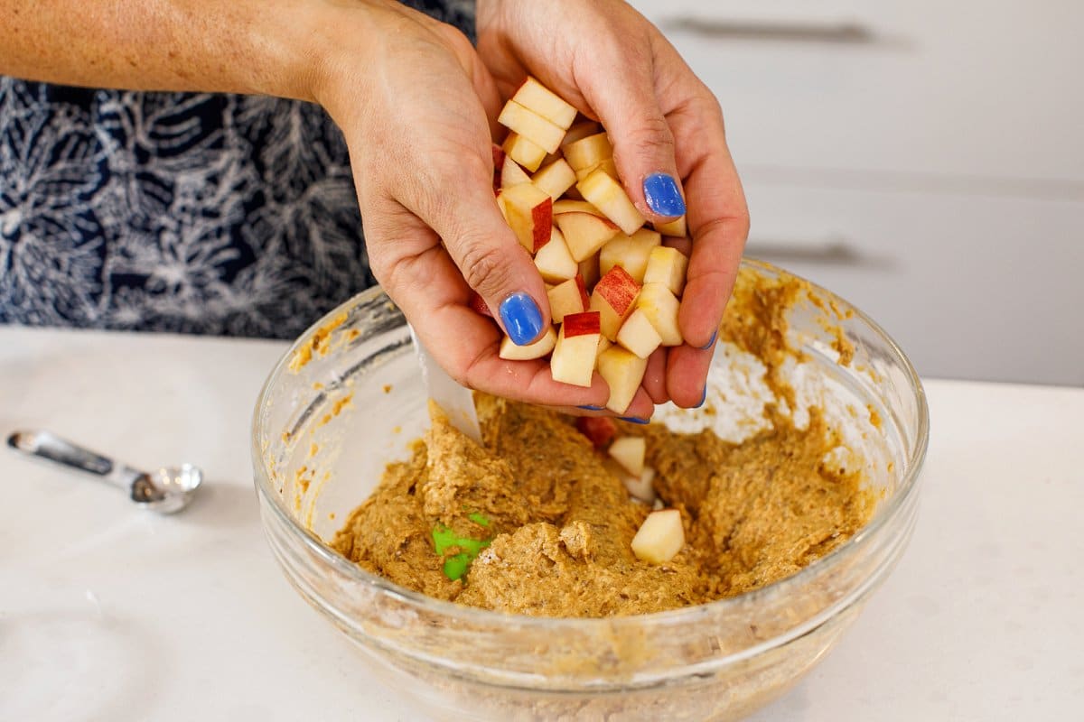 Liz's hands cupped full of chunks of apples being poured into the batter for pumpkin applesauce muffins in a glass bowl