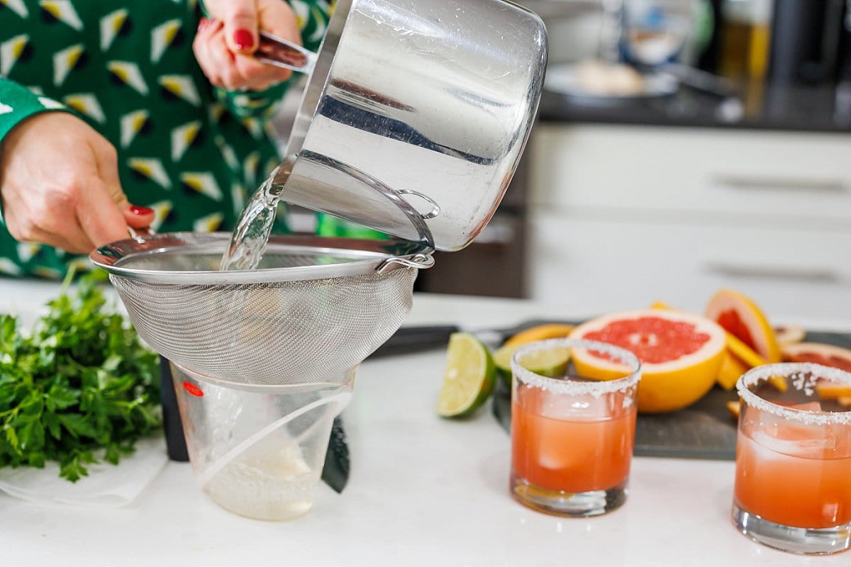 Straining rosemary simple syrup into a liquid measuring cup.