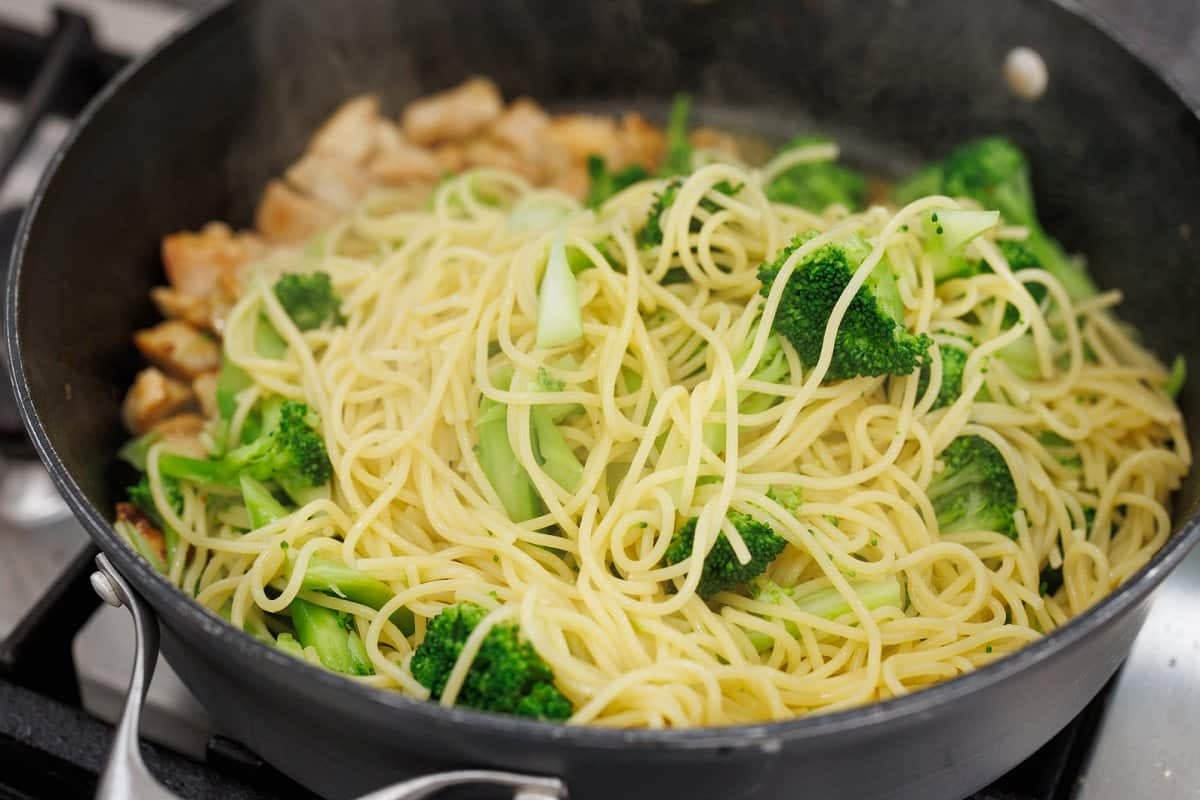 Cooked pasta and broccoli being added to pan with chicken.