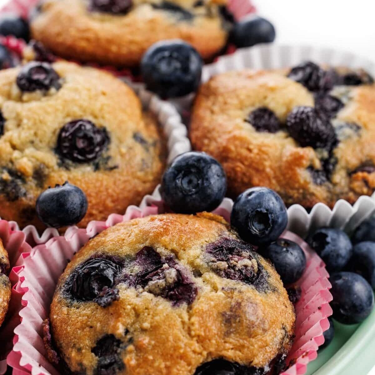 Gluten-free blueberry muffins in wrappers on a platter with fresh blueberries.