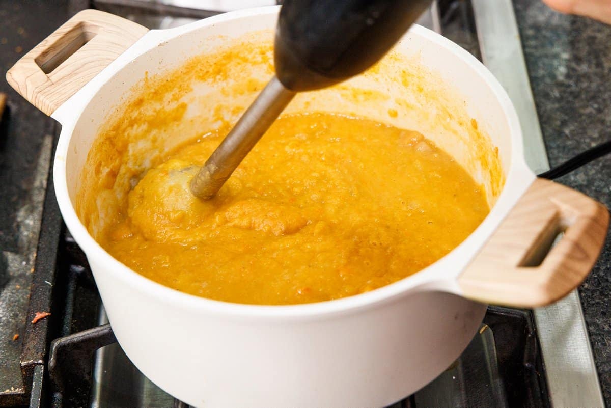Blending red lentil soup with an immersion blender in a white pot.